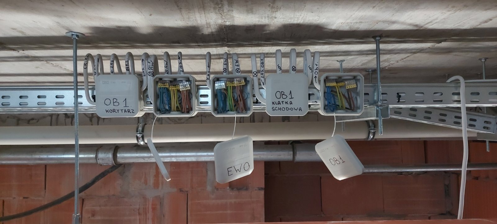 Rozprowadzenie przewodów elektrycznych w puszkach pod sufitem, widoczne kable w różnych kolorach, oznaczenia na puszkach: 'OB1 Korytarz', 'OB1 Klatka Schodowa', 'EWO', instalacja na etapie budowy...