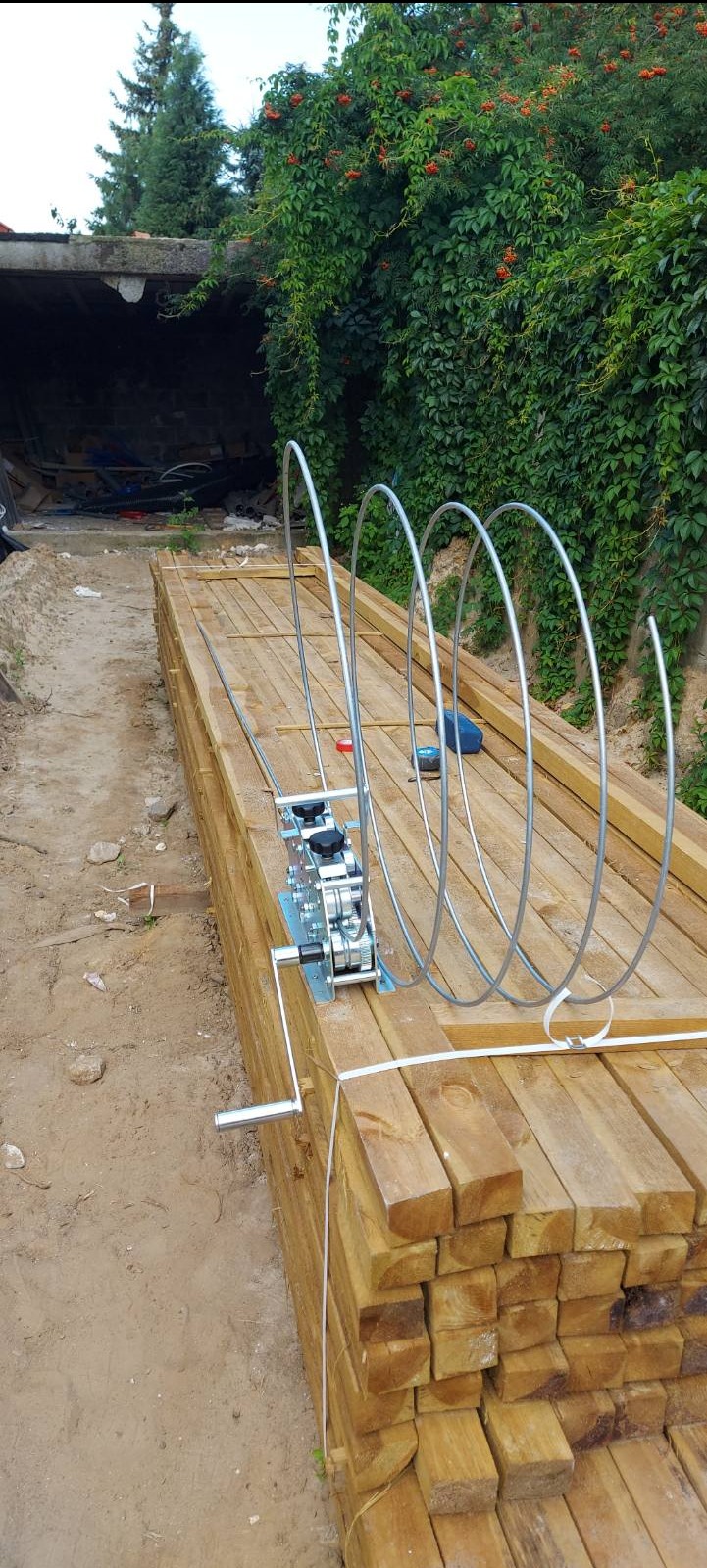 Niezamontowany, metalowy napęd do bramy na palecie drewnianych desek, widok z boku, zarośnięte tło.