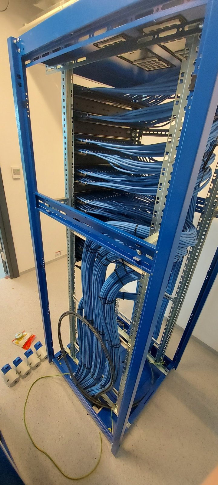 Niebieska szafa rack wypełniona uporządkowanymi kablami telekomunikacyjnymi, z zielonym kablem i złączkami na podłodze.