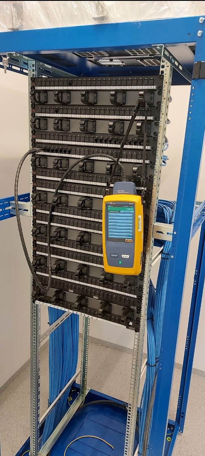 Panel krosowniczy w szafie rack, testowany żółtym miernikiem kabli, z widocznymi niebieskimi kablami sieciowymi.