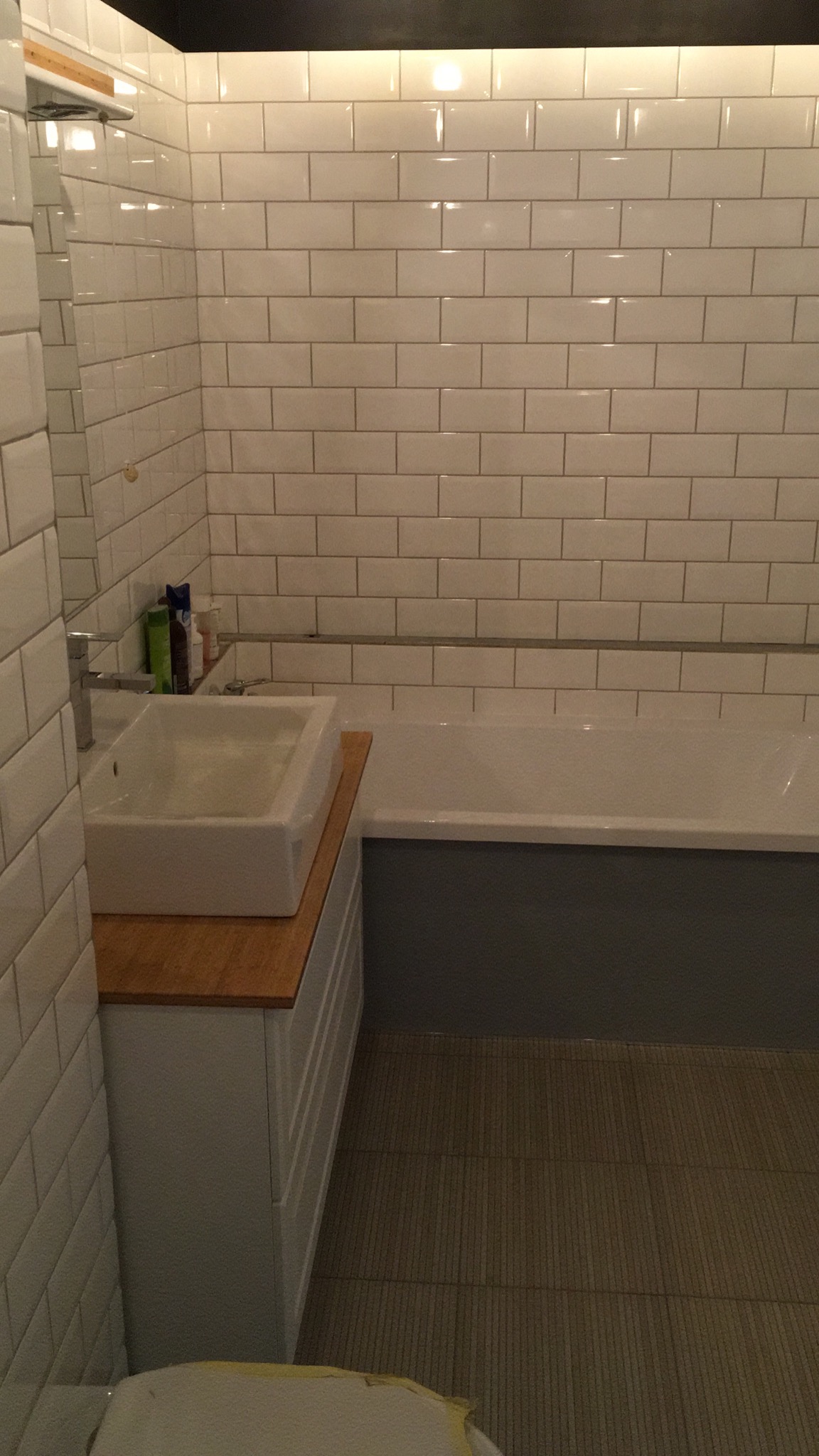 Wykończona łazienka z białymi, prostokątnymi płytkami na ścianach, białą umywalką nablatową na drewnianym blacie i szarą obudową wanny.