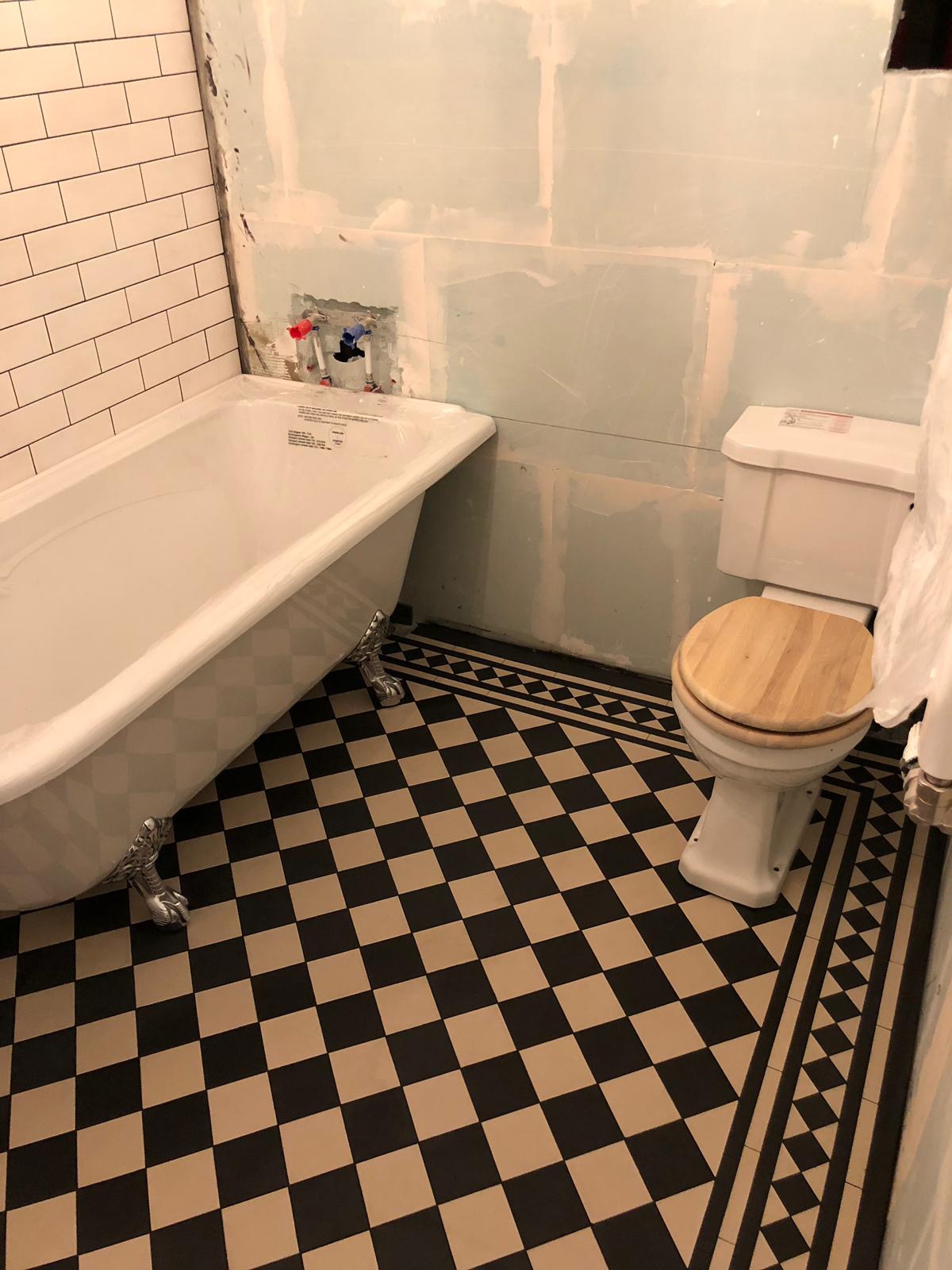Łazienka w trakcie remontu: wolnostojąca wanna na lwich nóżkach, toaleta z drewnianą deską, czarno-biała szachownica na podłodze, ściany częściowo wykończone płytkami, częściowo surowe płyty gipsowe.
