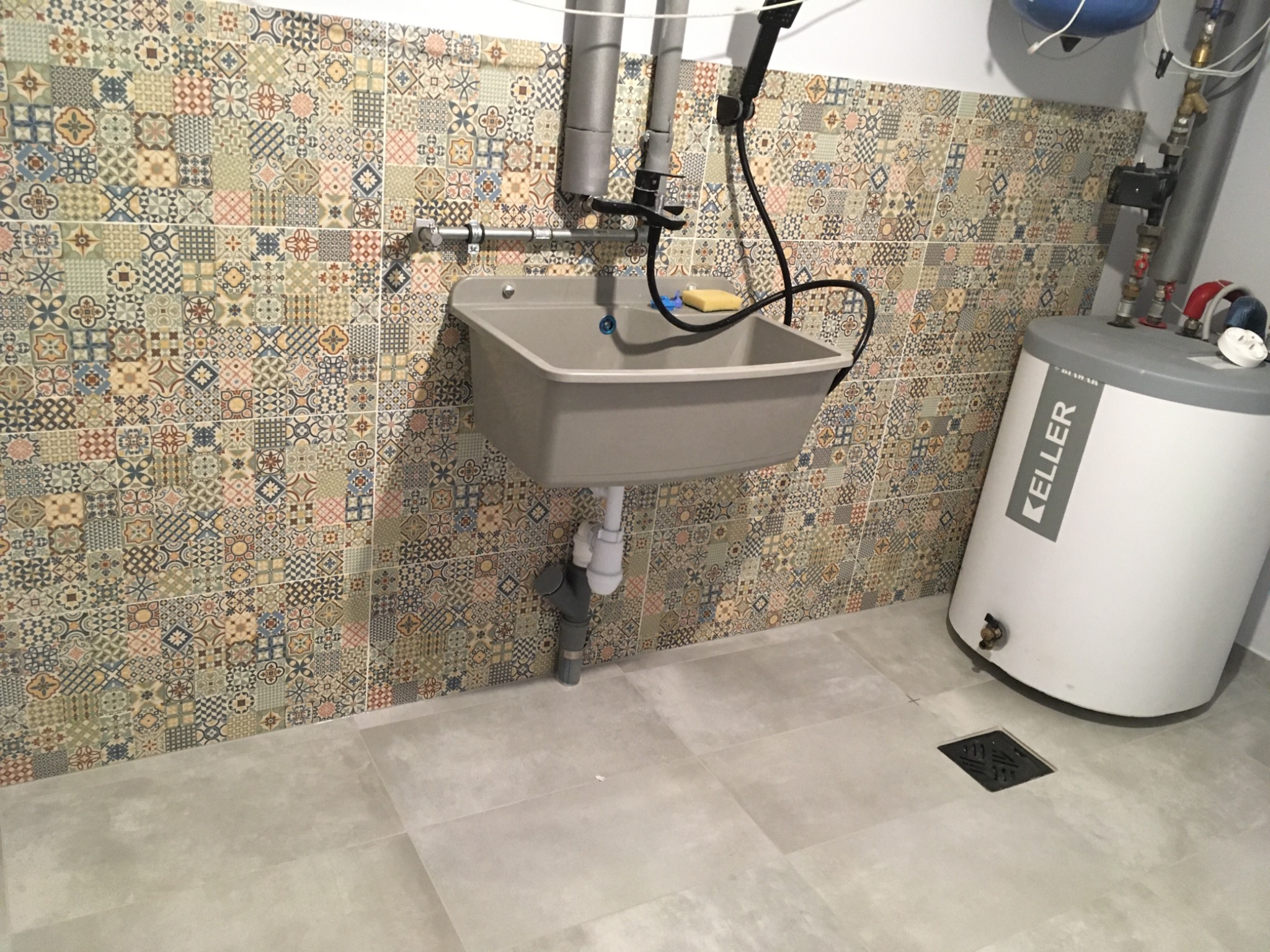 Szara umywalka gospodarcza zamontowana na ścianie z dekoracyjnymi, wzorzystymi płytkami, z widocznym bojlerem elektrycznym i odpływem podłogowym.
