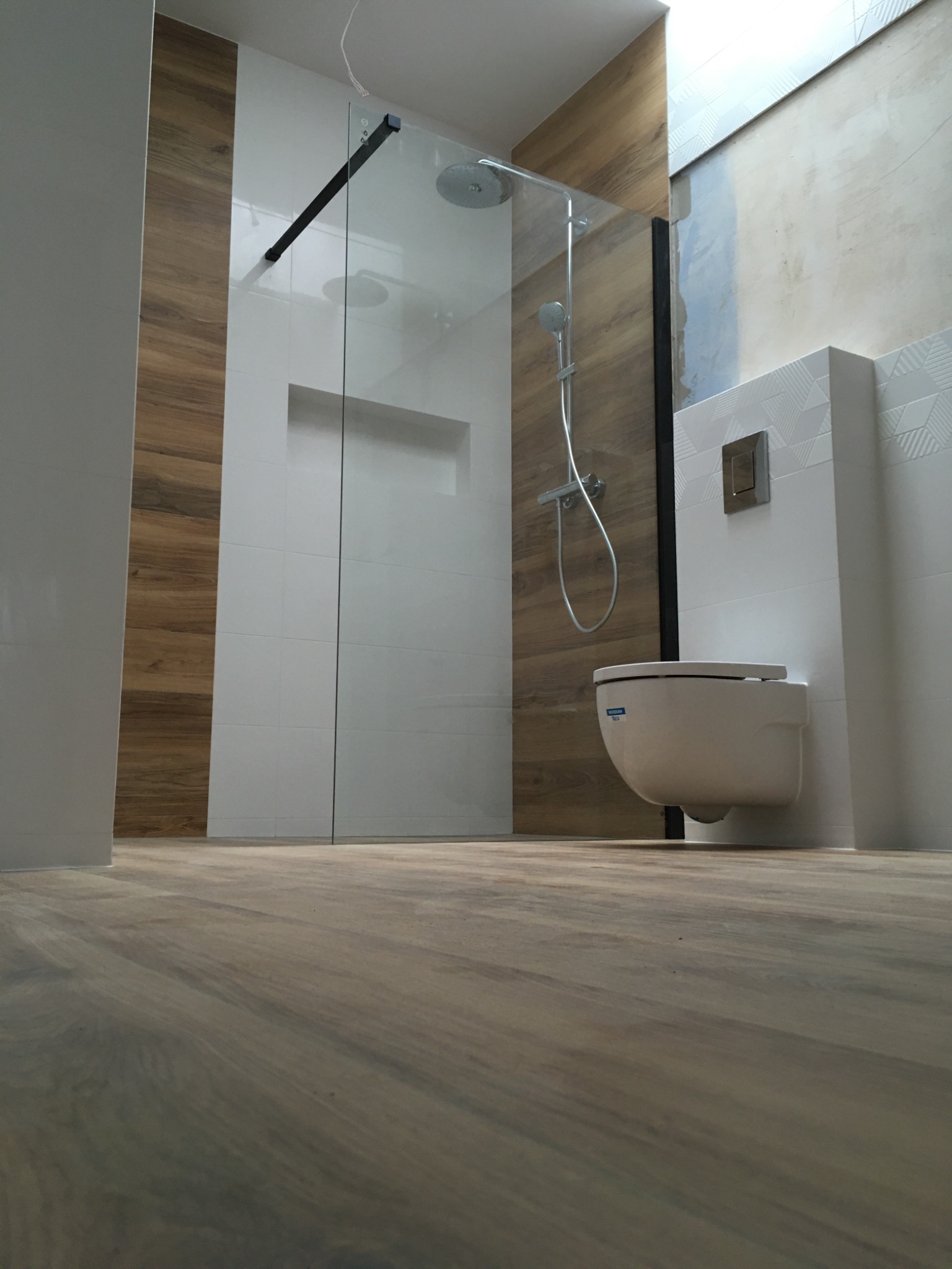 Wykończona łazienka w Lublinie z kabiną prysznicową typu walk-in z czarnymi elementami, wiszącą toaletą i drewnopodobnymi płytkami na ścianie oraz podłodze, widok z niskiej perspektywy.