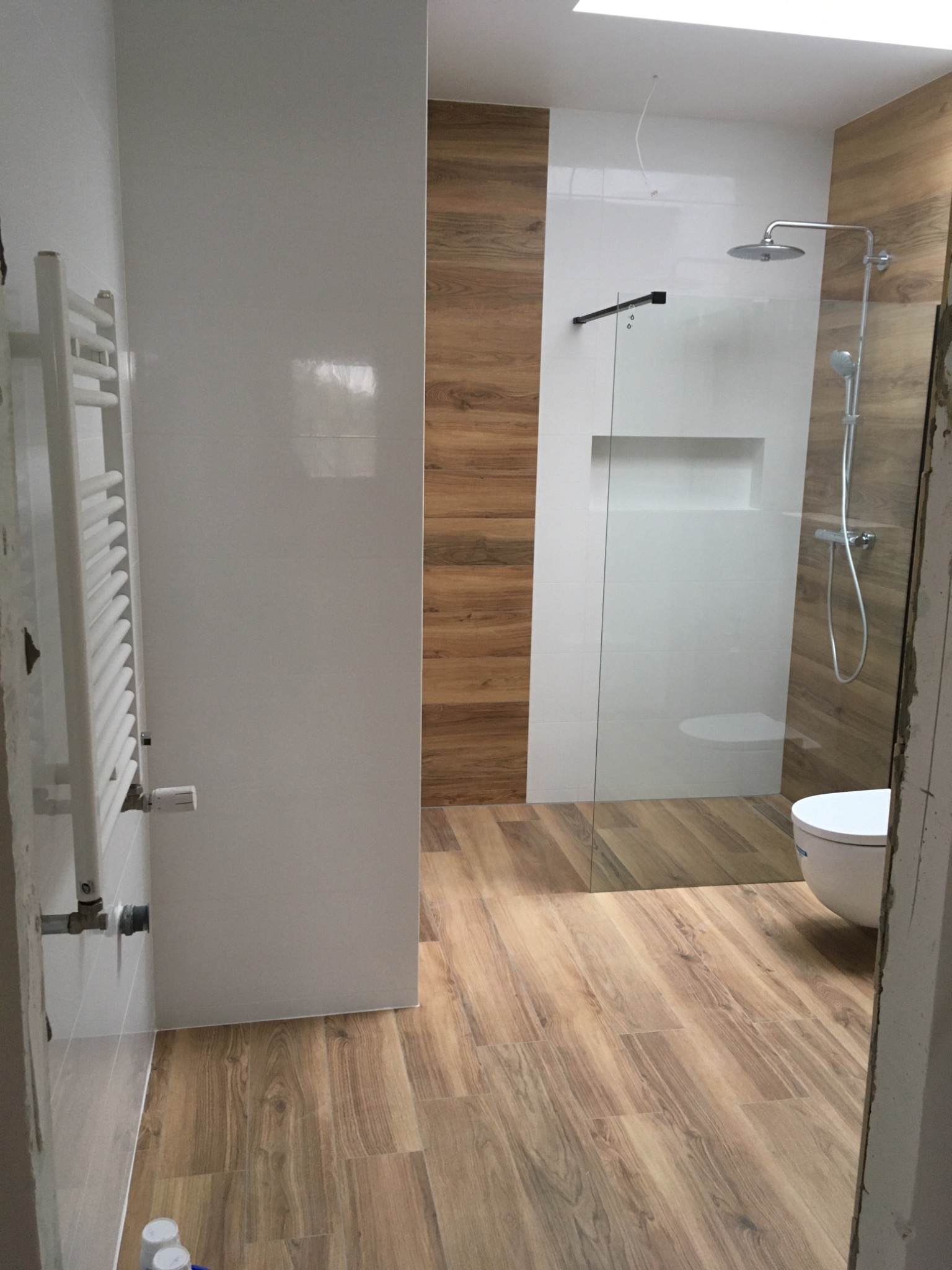 Wykończona łazienka z drewnopodobnymi płytkami na podłodze i ścianie prysznica, białe płytki na pozostałych ścianach, grzejnik drabinkowy po lewej, kabina prysznicowa ze szklaną ścianką...