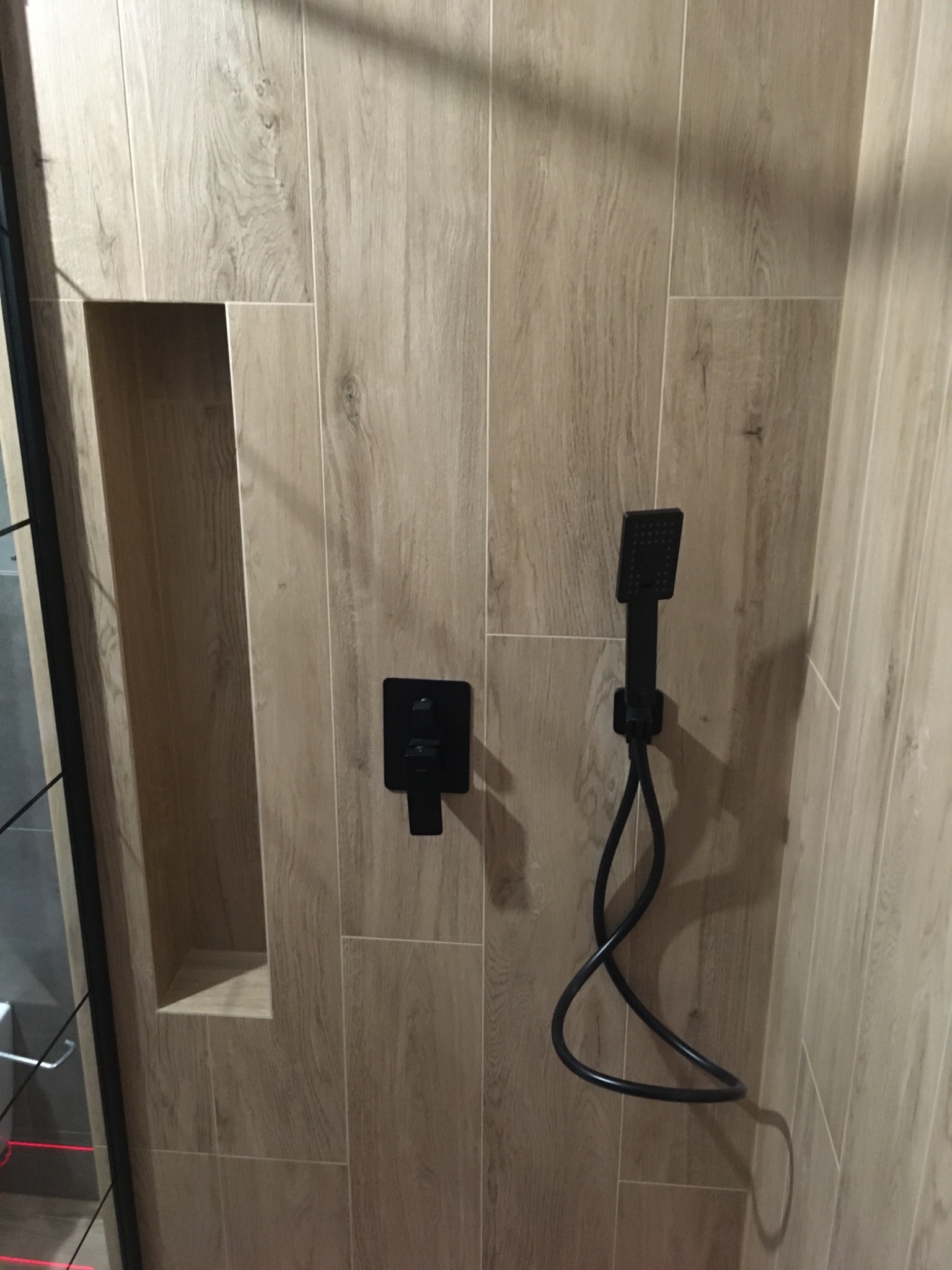 Wykończona ściana kabiny prysznicowej z beżowymi płytkami imitującymi drewno, wnęka w ścianie i czarna bateria z deszczownicą.