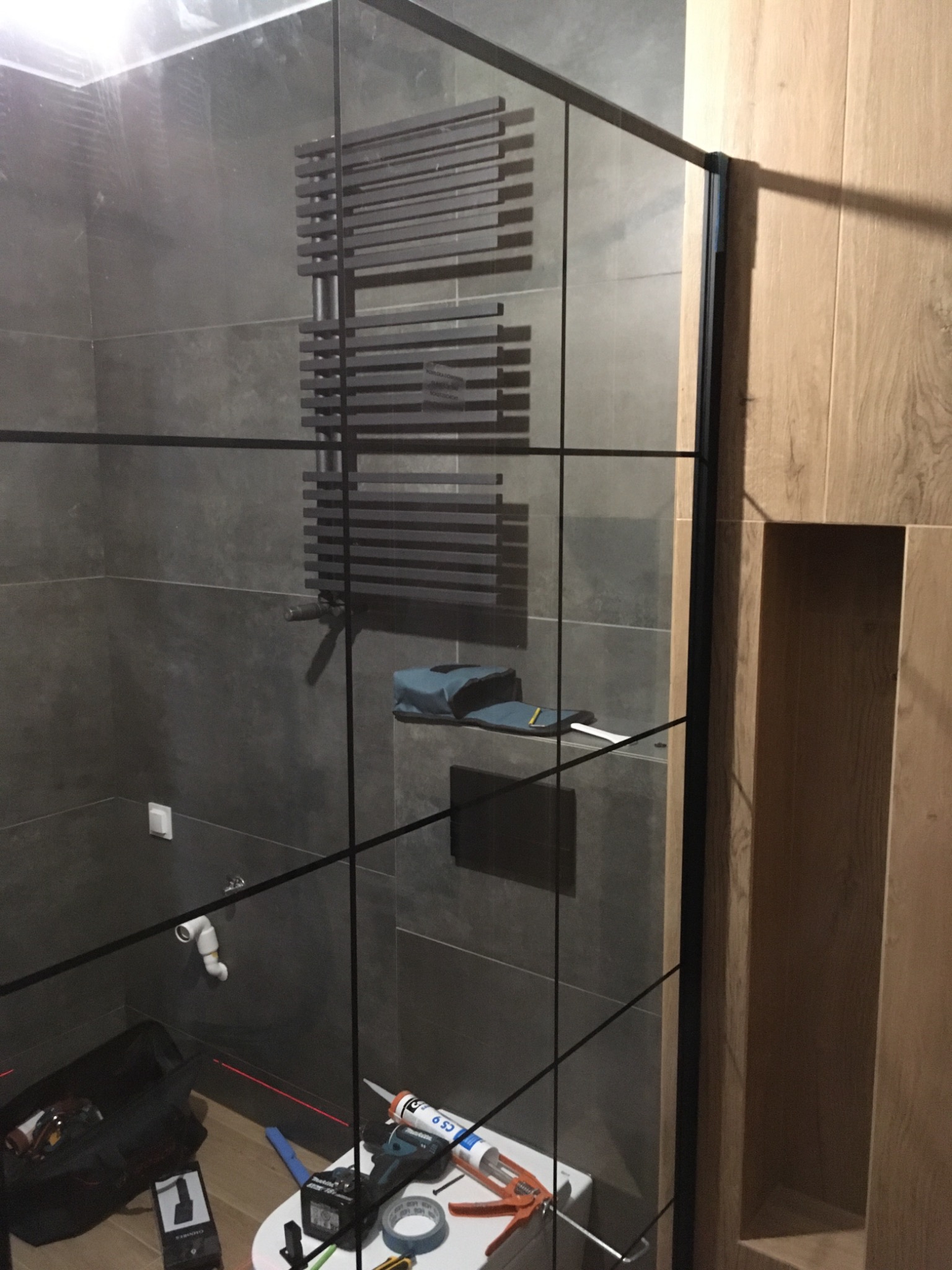 Szklana kabina prysznicowa z czarnymi profilami, widoczny grzejnik drabinkowy na szarej ścianie z płytek, narzędzia i akcesoria glazurnicze na podłodze, drewniana zabudowa z wnęką obok kabiny.