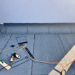 Fragment dachu pokrytego papą z narzędziami dekarskimi, widoczna butla gazowa, palnik, deska z narzędziami, rękawice robocze i metalowe pręty.