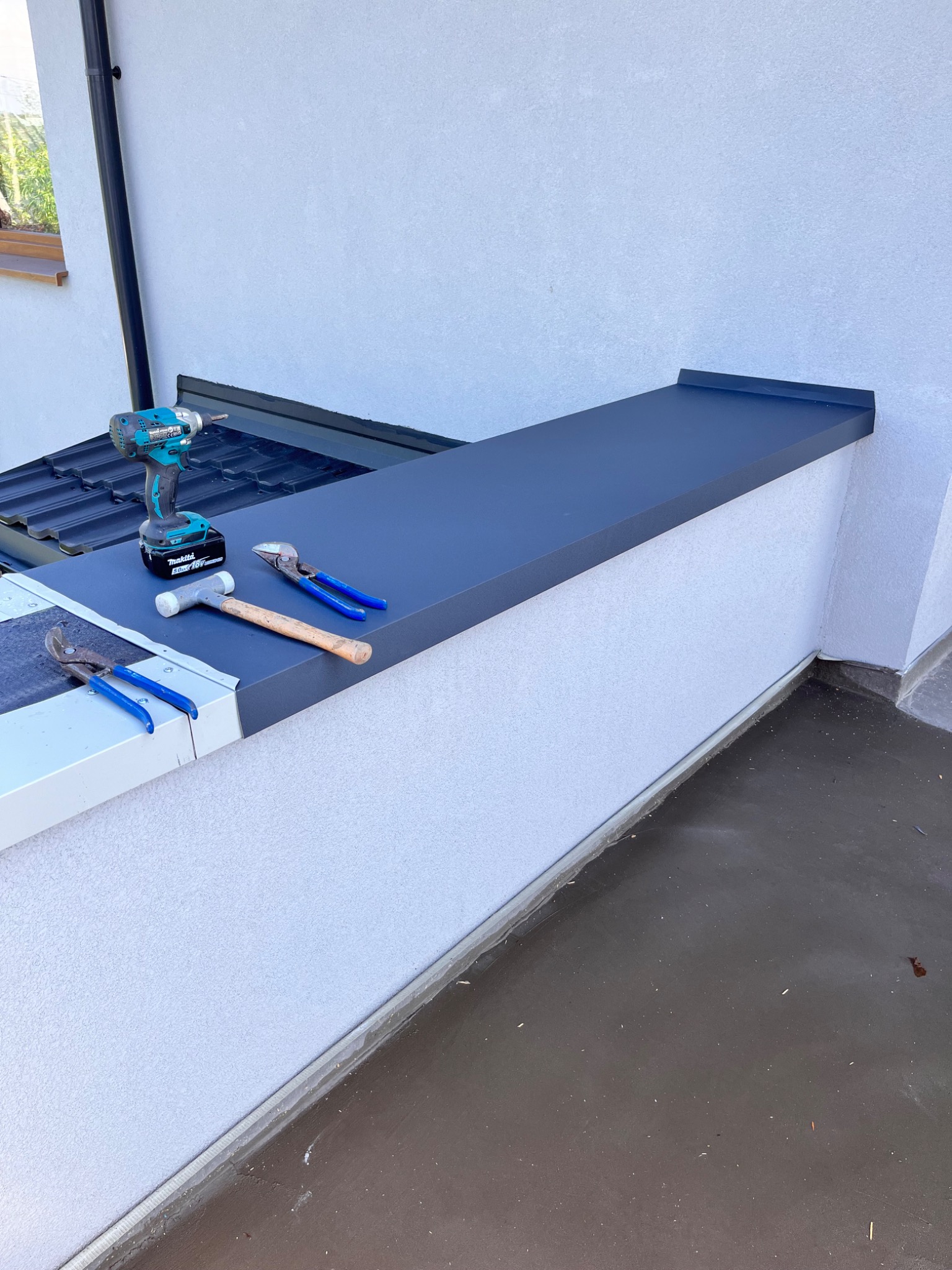 Szara, metalowa obróbka blacharska na dachu, widoczne narzędzia: wkrętarka akumulatorowa, młotek i dwa rodzaje szczypiec dekarskich.