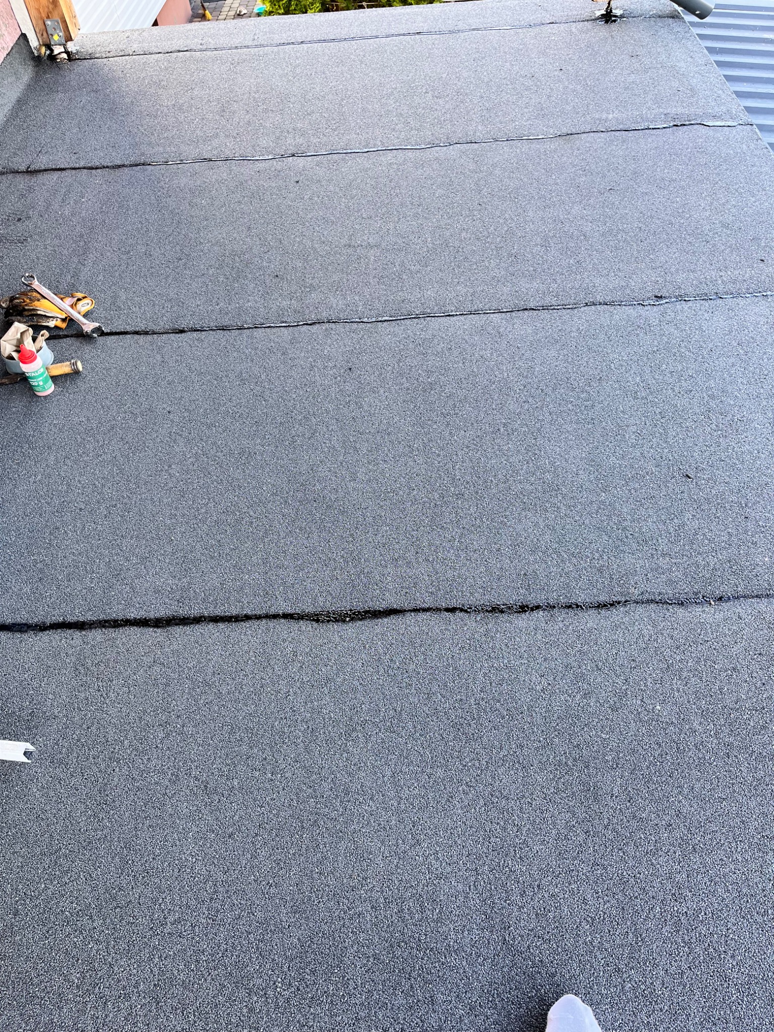 Czarny dach pokryty papą z widocznymi łączeniami, na lewo od środka narzędzia dekarskie i puszka z klejem, w tle fragment elewacji budynku.