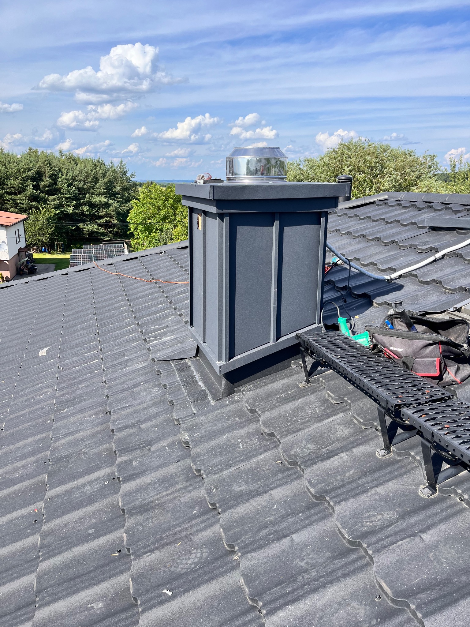 Szary dach z dachówki ceramicznej, obróbka komina z metalu w kolorze grafitowym, narzędzia dekarskie i drabinka na dachu, błękitne niebo z chmurami w tle.