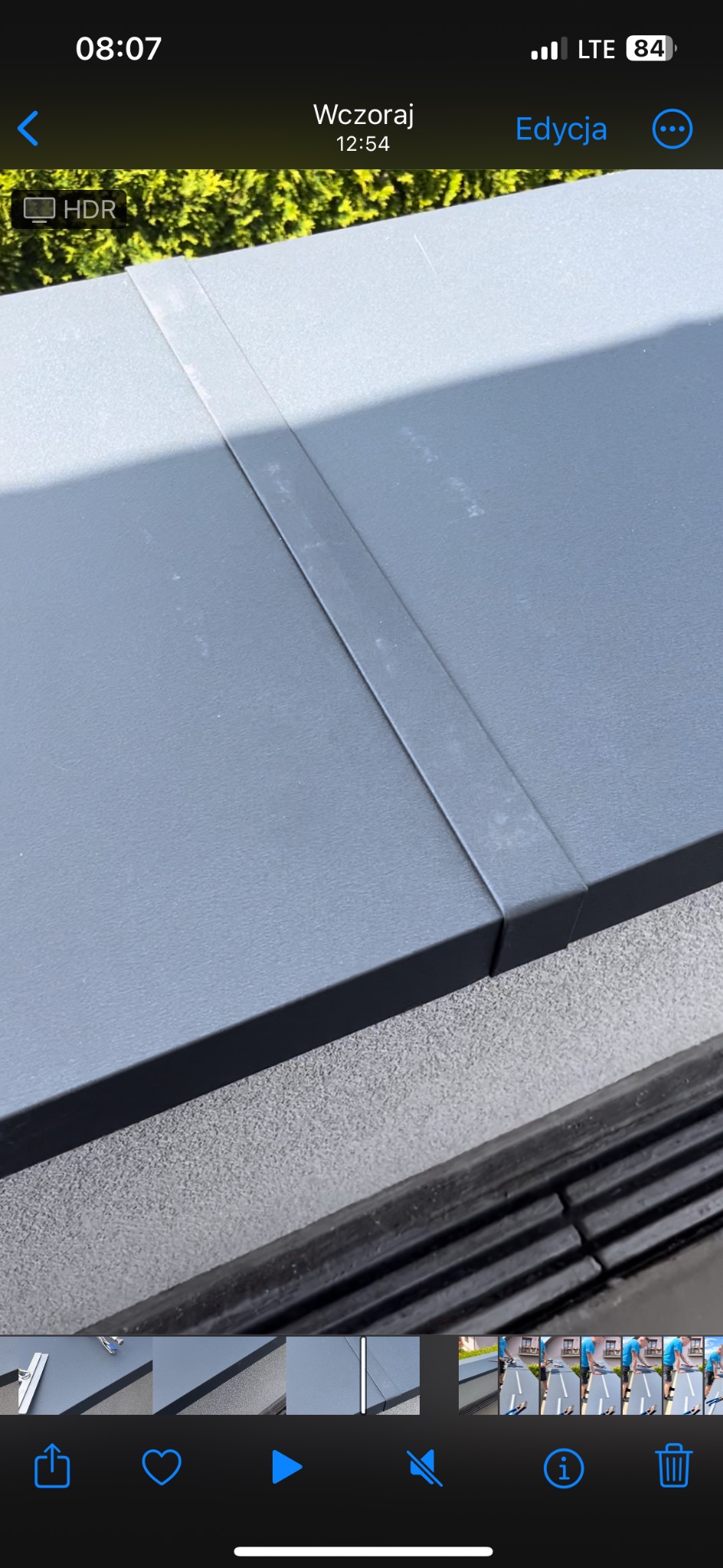 Szary dach z blachy trapezowej, widoczne połączenie arkuszy, zarys roślinności w tle.