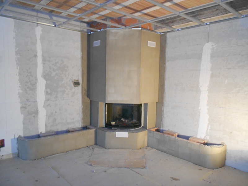 Wnętrze w trakcie budowy kominka narożnego z szarą obudową, widoczne niedokończone ściany, cegły i konstrukcja sufitu podwieszanego.