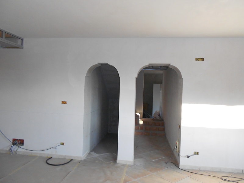 Wnętrze w trakcie remontu z dwoma łukowatymi przejściami, widoczne instalacje elektryczne, ślady po szpachlowaniu na ścianach i przygotowane podłoże pod podłogę.