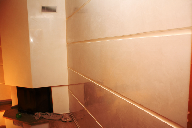 Ściana z dekoracyjnym stiukiem weneckim w ciepłych odcieniach beżu i brązu, podzielona poziomymi, podświetlanymi listwami, sąsiadująca z nowoczesnym kominkiem o białej obudowie.