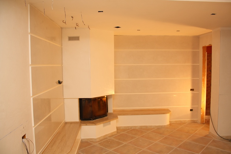 Salon w trakcie wykańczania z kominkiem narożnym, ściany zdobione stiukiem weneckim i poziomymi pasami, widoczne niedokończone prace elektryczne.