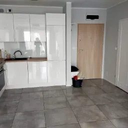 Nowoczesna, jasna kuchnia z błyszczącymi białymi szafkami, szarymi płytkami podłogowymi i drewnianymi blatami, widok na drzwi wejściowe i wewnętrzne, oraz wiadro z akcesoriami do sprzątania przy...