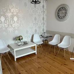 Poczekalnia z czterema białymi krzesłami Eames, białym stolikiem kawowym z kwiatami, zegarem ściennym i drewnianą podłogą, oświetlona lampą wiszącą w stylu loft