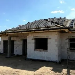 Dom w trakcie budowy, widoczny szkielet dachu pokryty częściowo czarną dachówką, ściany z białego betonu komórkowego, brak okien, w tle inny dom i błękitne niebo.