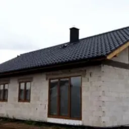 Dom jednorodzinny w stanie surowym zamkniętym z ciemnym, nowym dachem z dachówki ceramicznej i brązowymi oknami. Ściany z białego bloczka betonowego. Widoczny komin. Pochmurne niebo w tle.