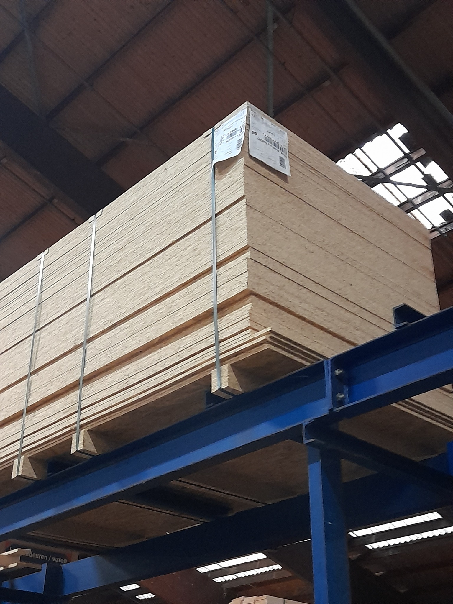 Stos płyt OSB zabezpieczonych metalowymi taśmami, umieszczony na niebieskiej stalowej konstrukcji w magazynie z widocznym dachem z świetlikami.
