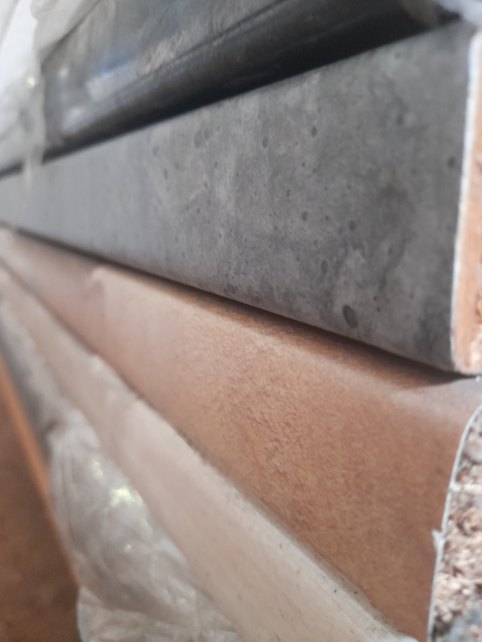 Stos drewnianych elementów wykończeniowych, prawdopodobnie listew przypodłogowych lub progowych, zabezpieczonych folią, z widoczną strukturą drewna i szarą, kamienną powłoką na niektórych elementach.