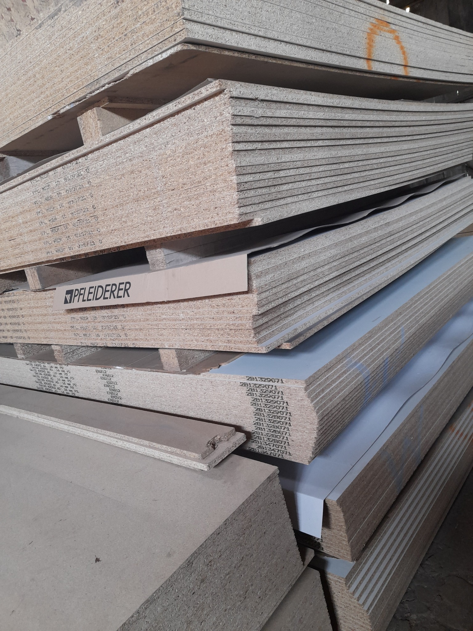 Stos płyt drewnopochodnych, widoczne płyty surowe i laminowane, ułożone na palecie, z widocznym logo producenta Pfleiderer.