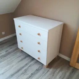 Biała komoda z czterema szufladami i okrągłymi, drewnianymi uchwytami, ustawiona na drewnianych nóżkach w pokoju ze skośnym sufitem i panelami podłogowymi imitującymi drewno.