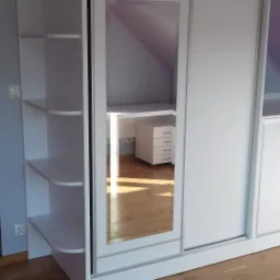 Biała szafa przesuwna z lustrem w pokoju na poddaszu, widoczne półki z zaokrąglonymi krawędziami i odbicie biurka z szufladami.