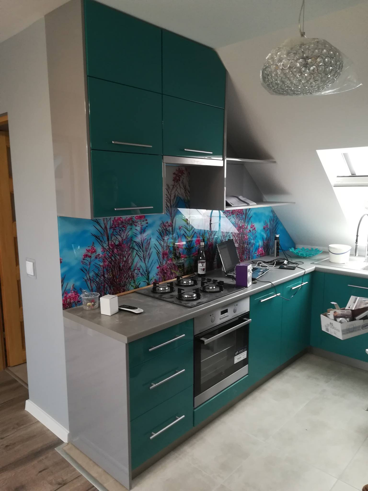Nowoczesna kuchnia z turkusowymi szafkami na wysoki połysk, szarym blatem i panelem szklanym z motywem kwiatowym, oświetlona kryształowym żyrandolem w folii.