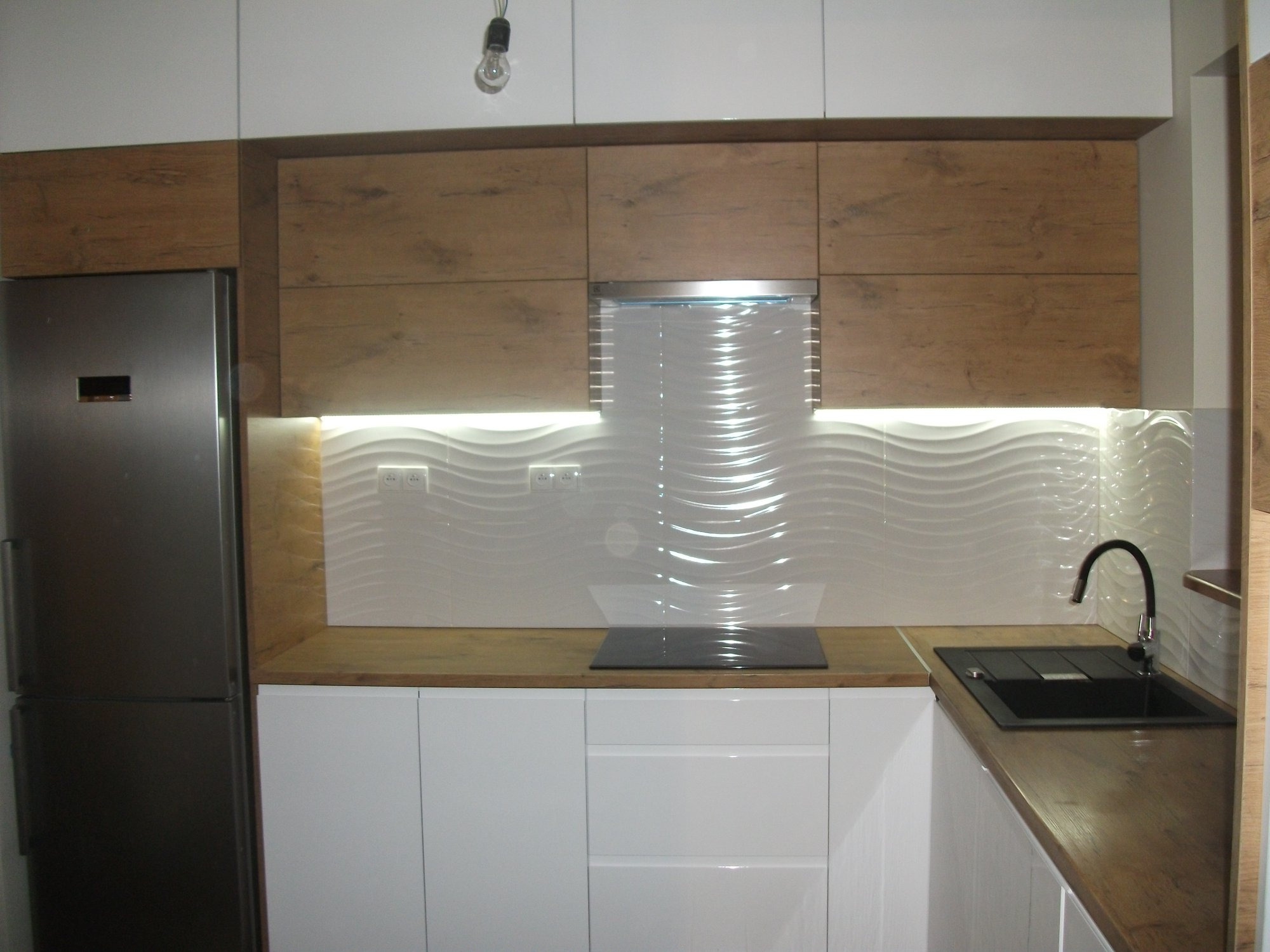 Nowoczesna kuchnia z białymi frontami i drewnianymi blatami, faliste płytki na ścianie, srebrna lodówka po lewej stronie, czarny zlew i bateria po prawej, widoczna płyta indukcyjna.