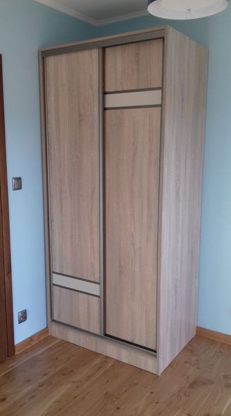 Jasna, drewnopodobna szafa wnękowa z przesuwnymi drzwiami, z poziomymi wstawkami w kolorze szarości, na tle błękitnej ściany i drewnianej podłogi.