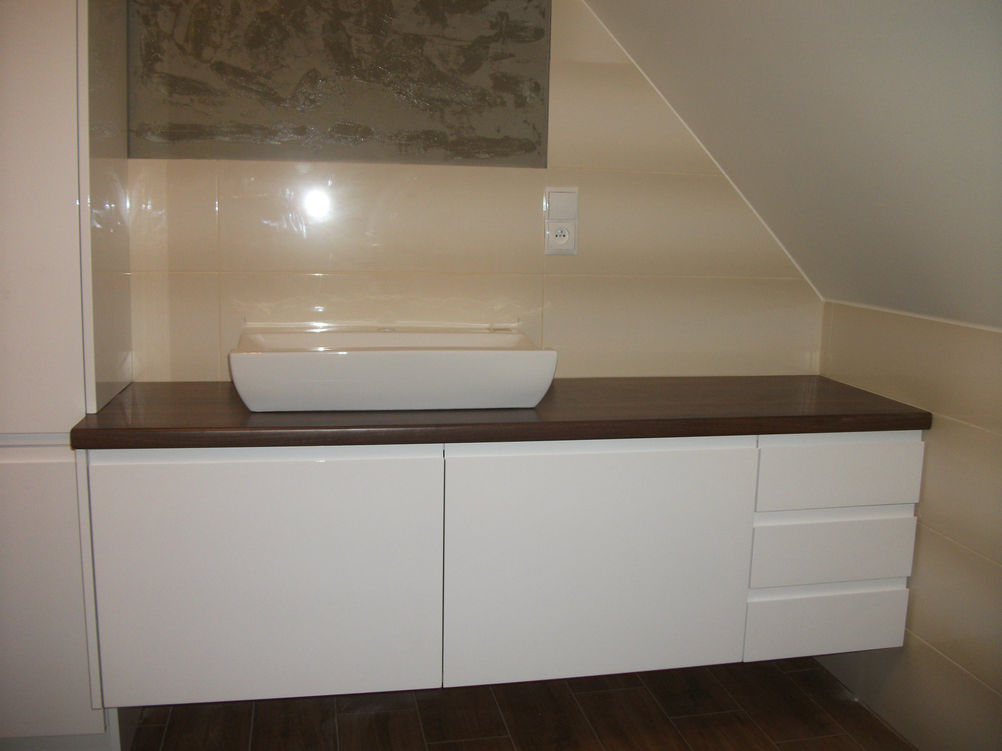 Biała szafka łazienkowa pod umywalkę z ciemnym blatem drewnianym w skosie poddasza, widoczne trzy szuflady po prawej stronie.