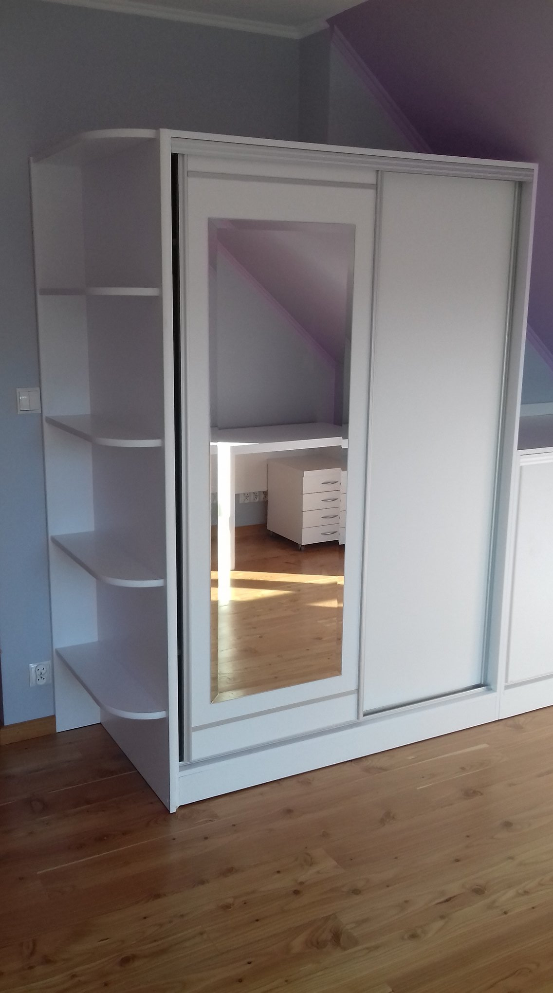 Biała szafa przesuwna z lustrem w pokoju na poddaszu, widoczne półki z zaokrąglonymi krawędziami i odbicie biurka z szufladami.