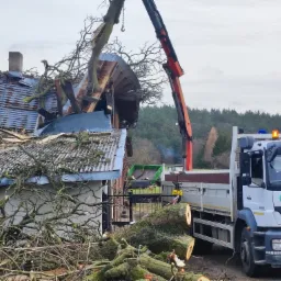 Uszkodzony budynek po upadku drzewa, fragment drzewa podnoszony dźwigiem, w tle ciężarówka z otwartą skrzynią ładunkową.
