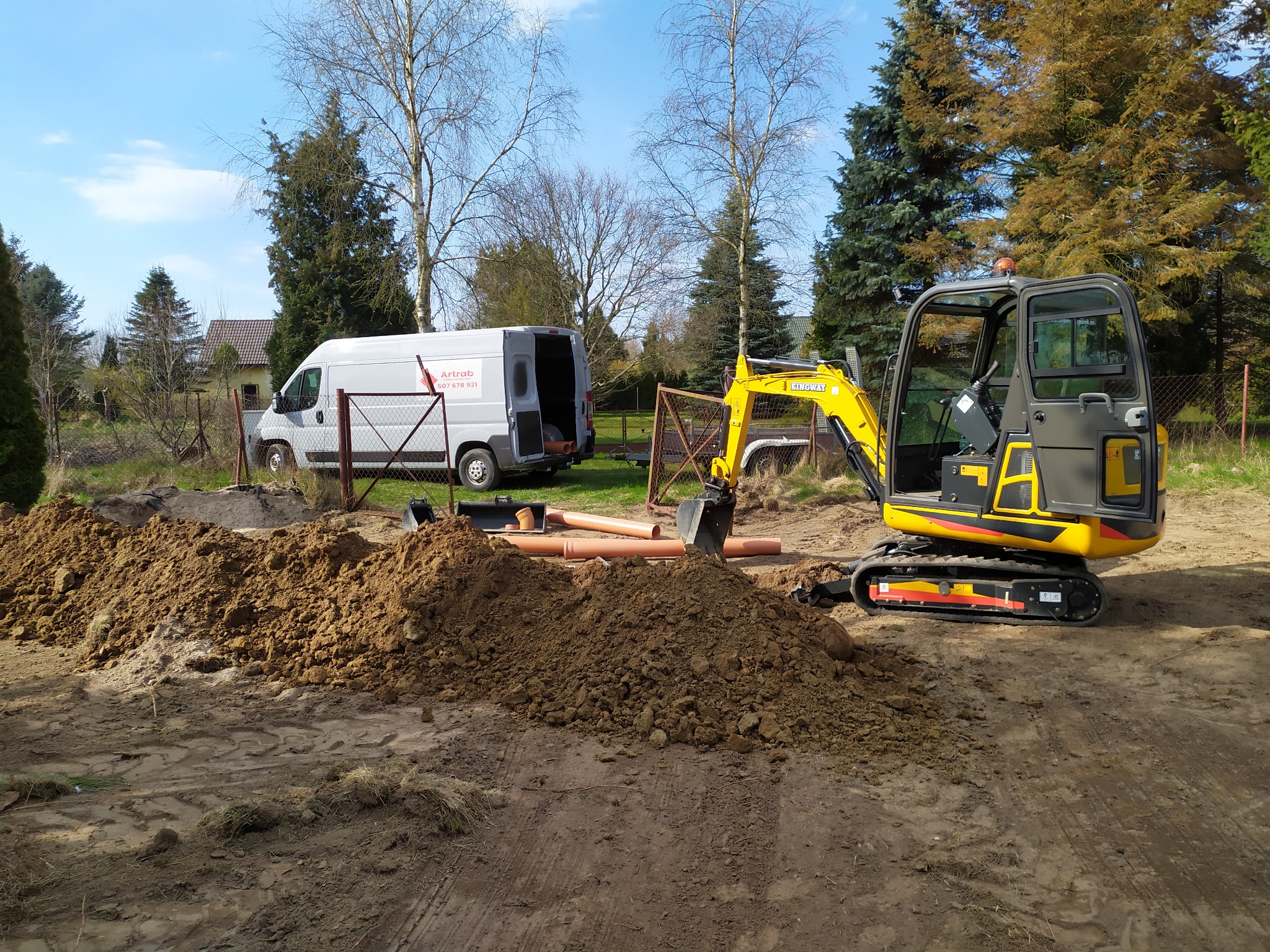 Żółta mini koparka Kingway na placu budowy, obok sterta ziemi, rury kanalizacyjne i biały bus firmy ArtRAB, w tle dom i drzewa.