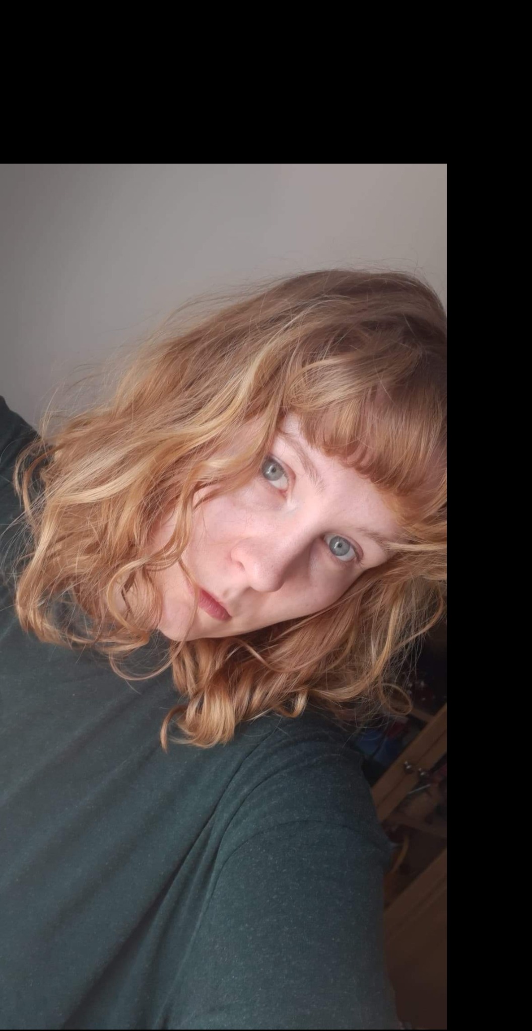 Portret kobiety z falowanymi rudymi włosami i grzywką, ubrana w ciemnozieloną koszulkę, spoglądająca w dół z lekkim uśmiechem.