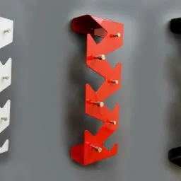 Trzy metalowe wieszaki na ubrania w kolorach białym, czerwonym i czarnym, zamontowane na szarej ścianie, z charakterystycznym zygzakowatym kształtem i wystającymi kołkami.