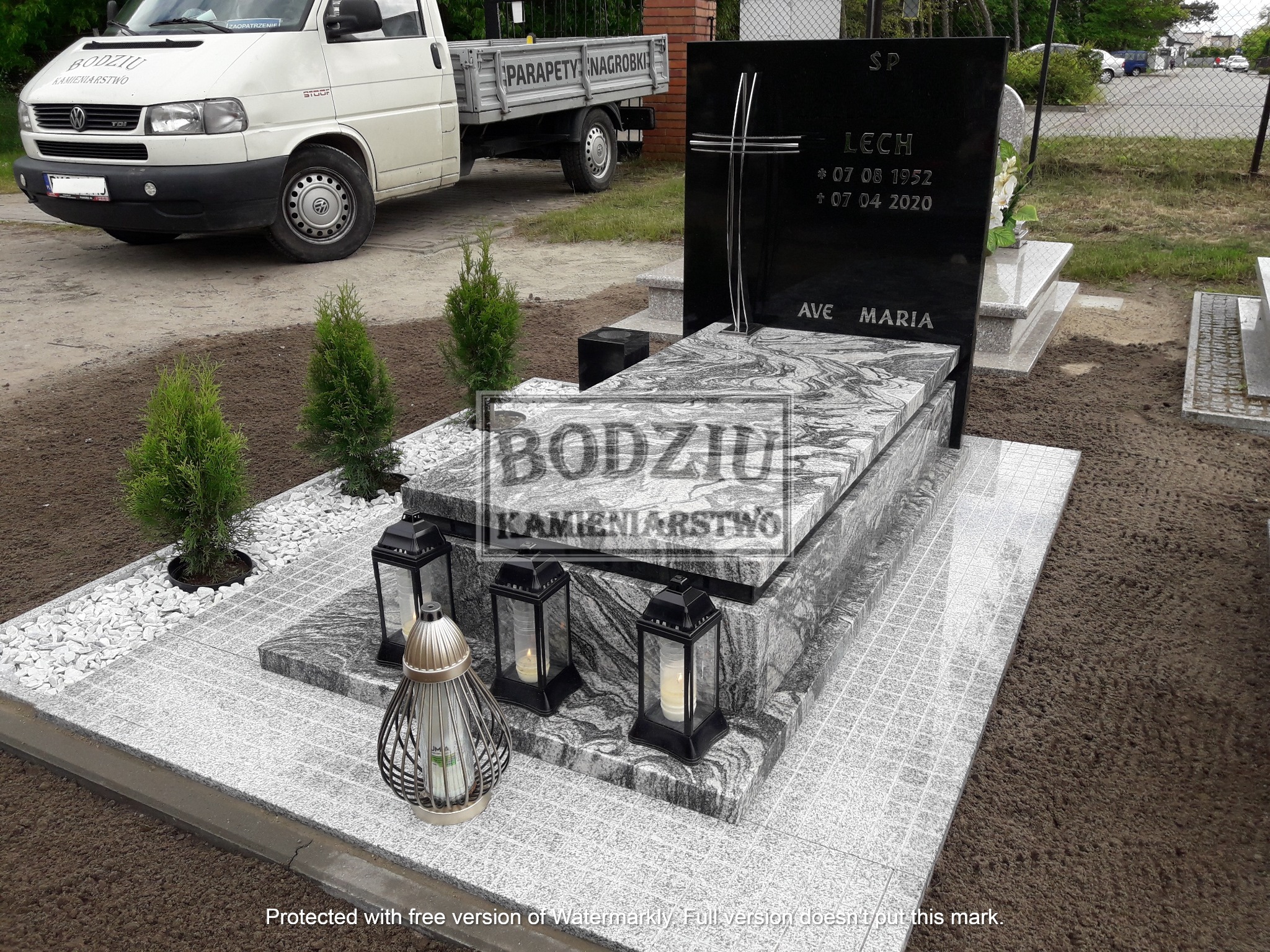 Granitowy nagrobek z czarną tablicą inskrypcyjną, ozdobiony krzyżem i napisem 'Ave Maria', otoczony lampionami i małymi tujami, na tle firmowego samochodu dostawczego.