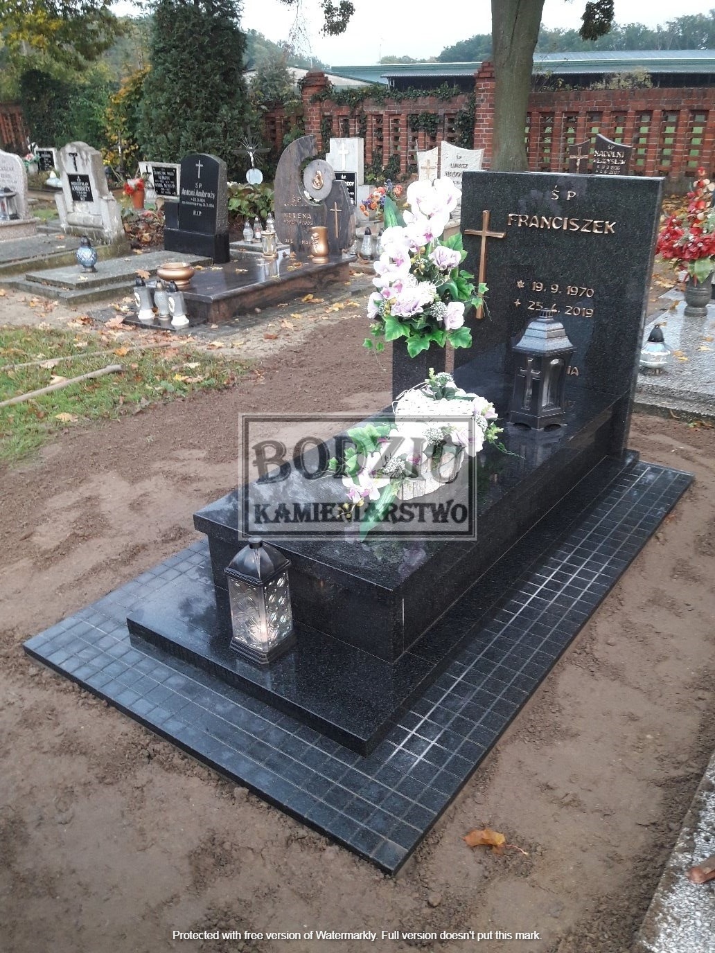 Czarny, granitowy nagrobek z imieniem Franciszek, datami urodzenia i śmierci, ozdobiony kwiatami i lampionem, na cmentarzu, z widocznym logo firmy kamieniarskiej.