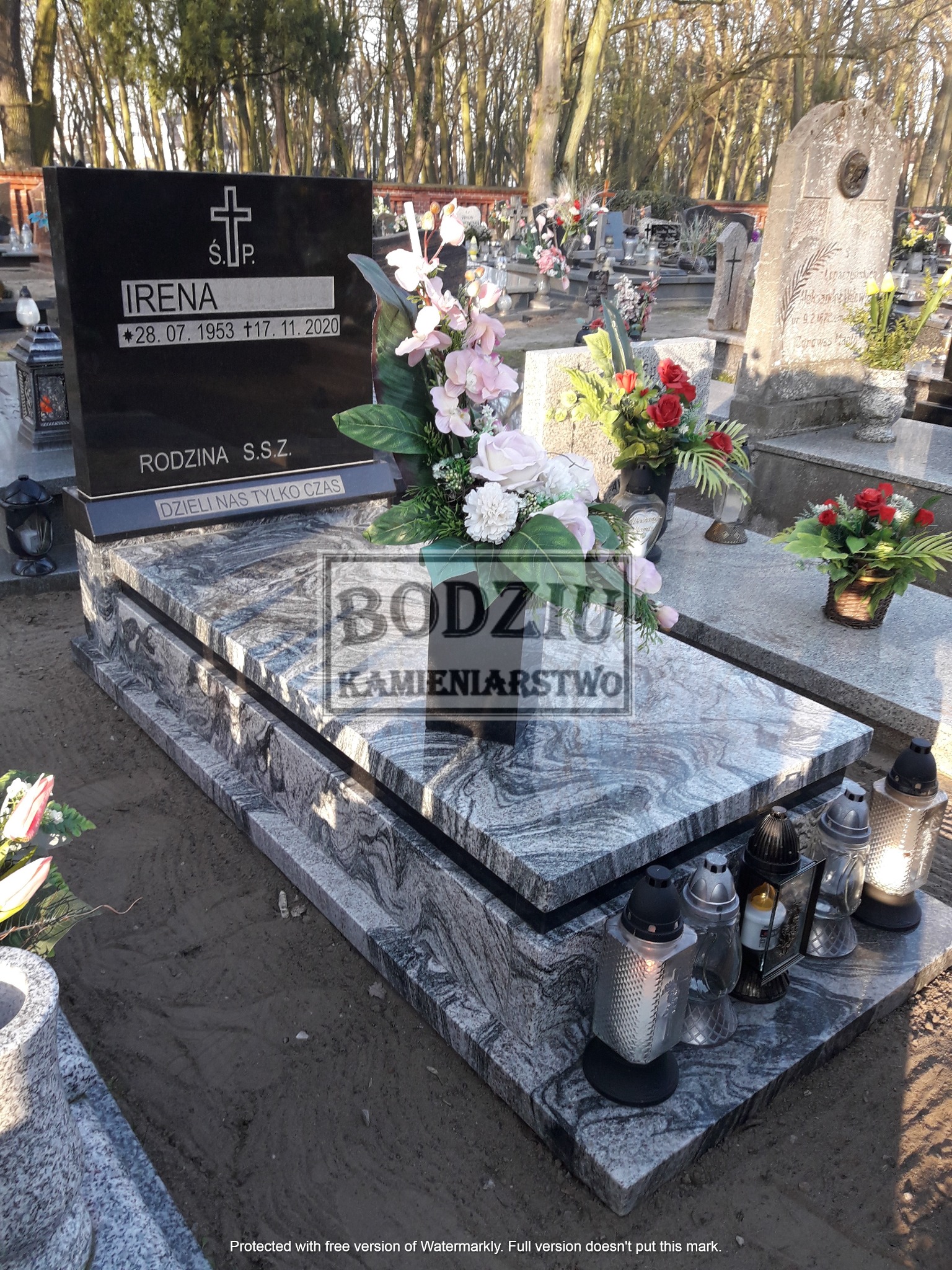 Czarny nagrobek z jasnoszarego kamienia z datami urodzin i śmierci Ireny, ozdobiony kwiatami i zniczami, na tle cmentarza.