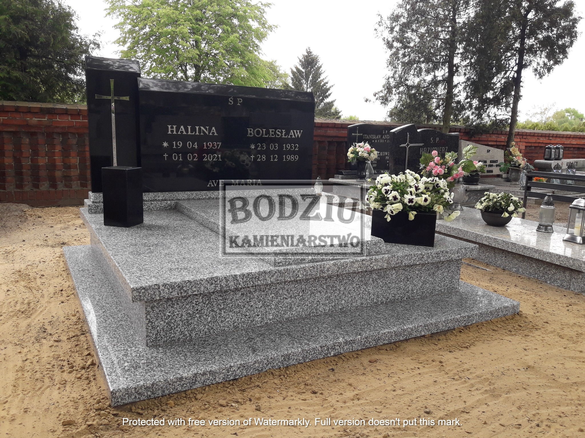 Czarny, granitowy nagrobek z metalowym krzyżem i inskrypcjami na dwupoziomowej, szarej podstawie, udekorowany kwiatami w czarnych donicach.