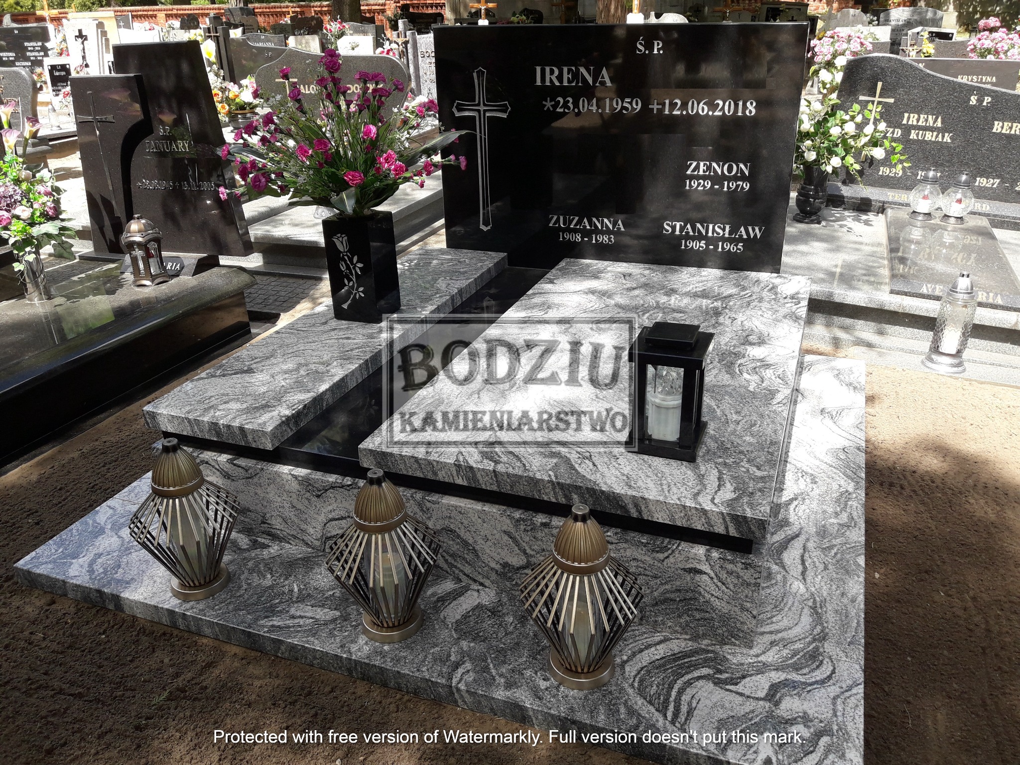 Granitowy nagrobek z czarną tablicą, wazonem z różowymi kwiatami i trzema lampionami w metalowych obudowach na cmentarzu.