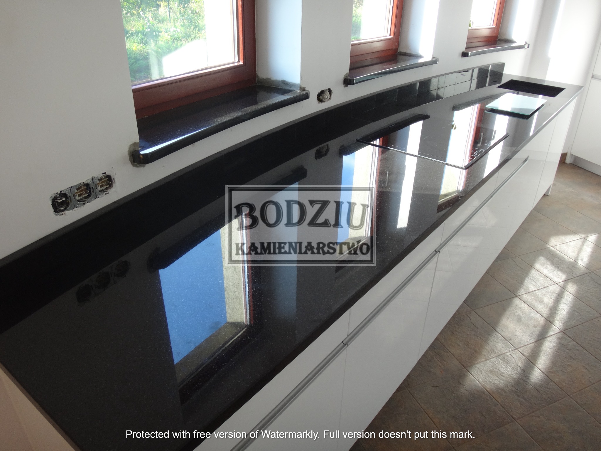 Czarny kamienny blat kuchenny z widocznymi gniazdkami elektrycznymi i oknami, błyszczący odbijający światło, z logo firmy kamieniarskiej w centralnej części blatu.