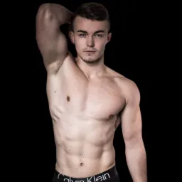 Młody mężczyzna z krótkimi włosami, bez koszulki, prezentuje umięśnioną sylwetkę na czarnym tle, z ręką uniesioną nad głową.