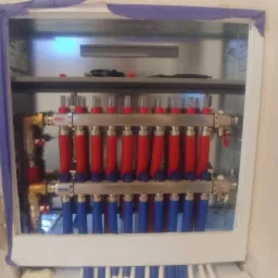 Rozdzielacz ogrzewania podłogowego z czerwonymi i niebieskimi rurami w metalowej szafce, widoczny fragment instalacji w podłodze i nieotynkowana ściana z niebieską taśmą malarską.