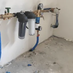 Zestaw filtrów wody zamontowany na ścianie, widoczne rury doprowadzające i odprowadzające wodę, manometry oraz zawory odcinające. Surowy stan posadzki.