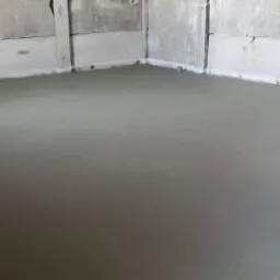 Świeżo wylana, gładka posadzka w szarym odcieniu, widoczne surowe ściany z betonu komórkowego i fragment futryny drzwi po lewej stronie.