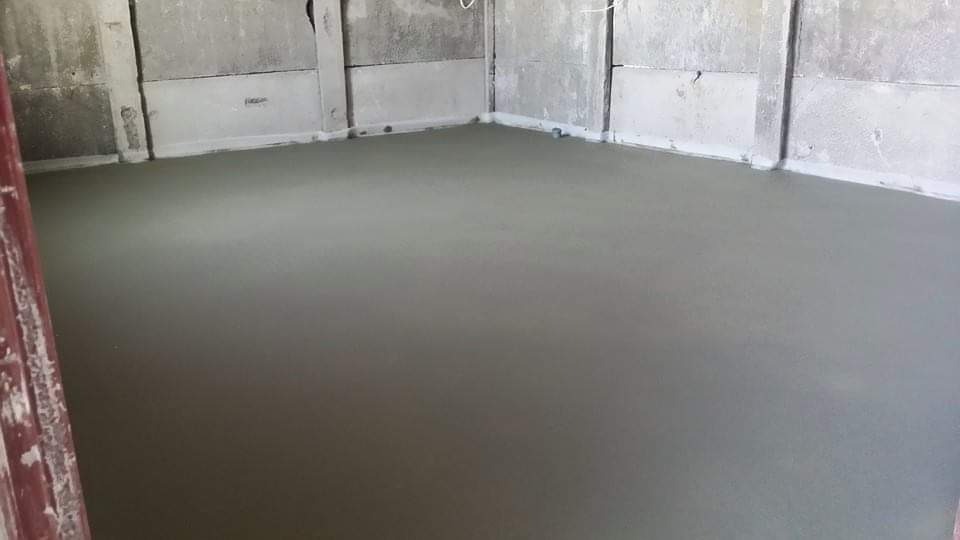 Świeżo wylana, gładka posadzka w szarym odcieniu, widoczne surowe ściany z betonu komórkowego i fragment futryny drzwi po lewej stronie.