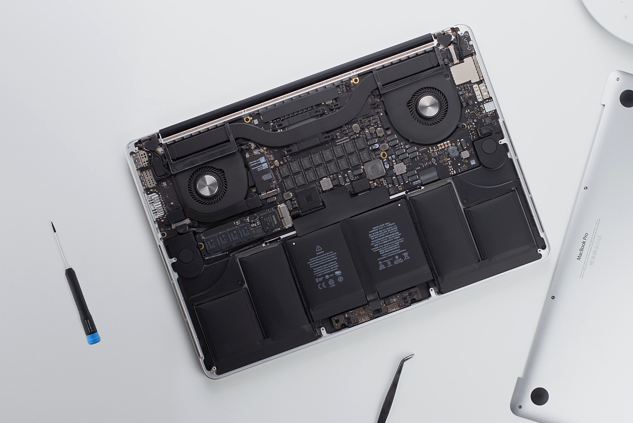 Wnętrze otwartego laptopa MacBook Pro z widoczną płytą główną, baterią i wentylatorami, obok leżą śrubokręt i pęseta na białym tle.